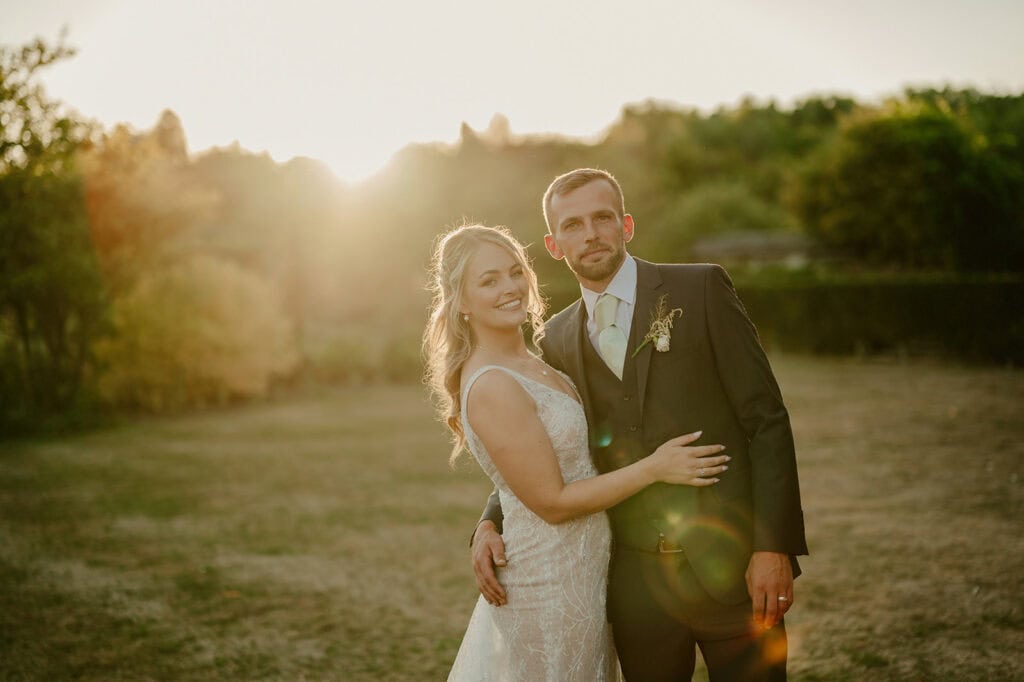 A couple dressed in wedding attire stands outdoors in a sunlit field, with the woman in a white gown and the man in a dark suit. They are embracing and looking at the camera while smiling gently.