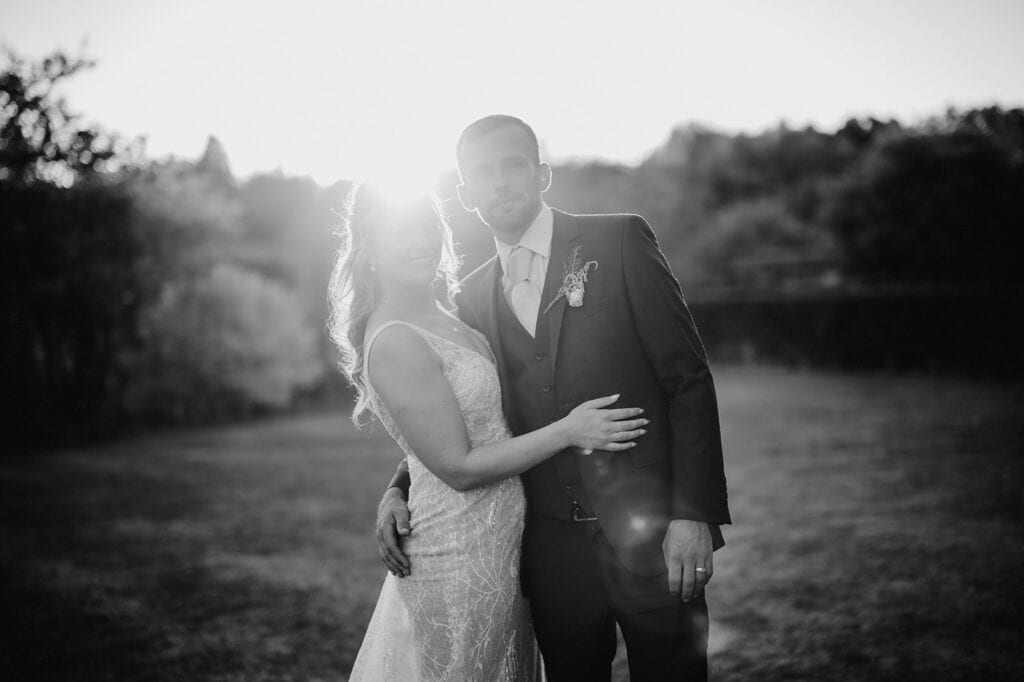 A bride and groom stand outdoors embracing, with the sun setting in the background. The photo is in black and white.