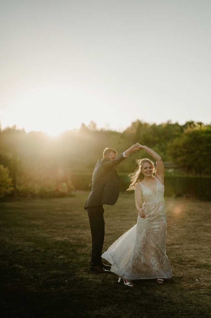 A couple dressed in wedding attire dance outdoors on a grassy area at sunset, with the sun casting a warm glow behind them.