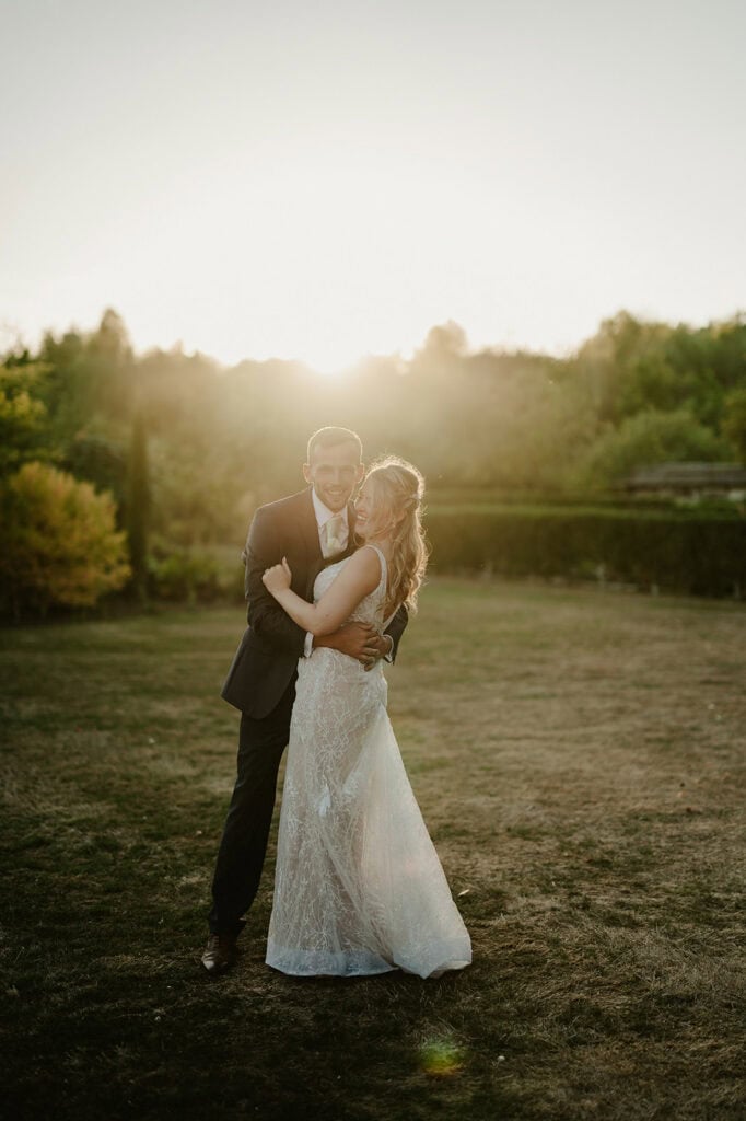 A couple stands together in an outdoor setting at sunset, smiling and embracing. The woman wears a lace wedding dress; the man is dressed in a suit. Trees are visible in the background.
