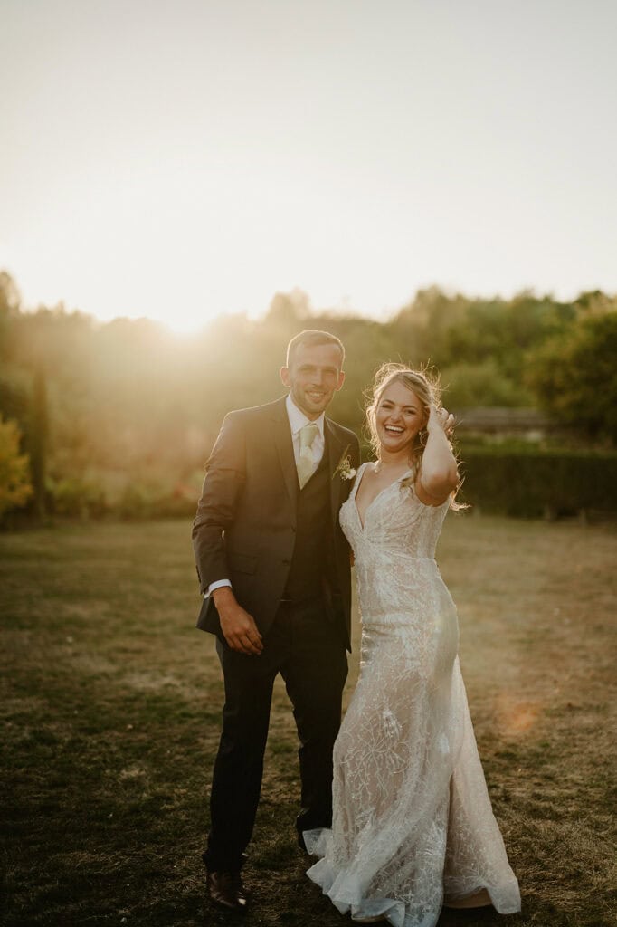 A couple in wedding attire stands outdoors on a sunny day, smiling and looking towards the camera. The bride wears a white dress, and the groom is in a suit. The setting includes green trees and a grassy area.