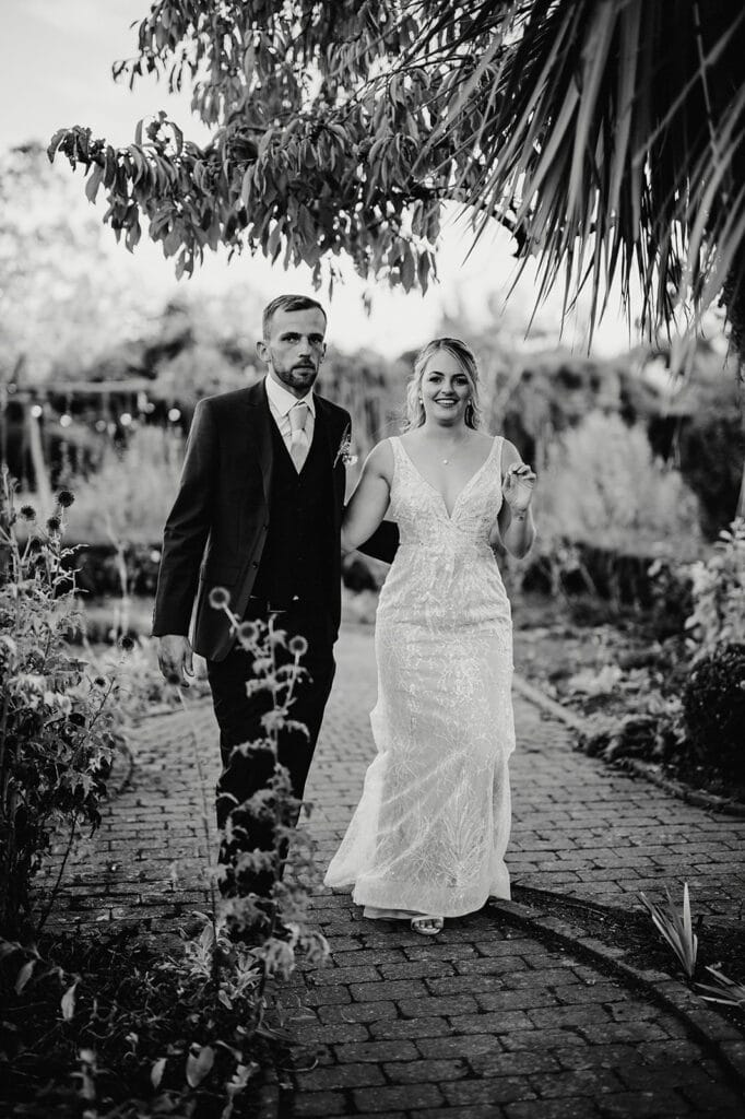 A bride and groom walk hand-in-hand down a brick path in an outdoor garden setting. The bride wears a lace gown, and the groom is in a dark suit. Both are looking slightly forward.