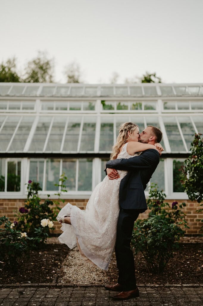 A groom lifts and embraces a bride in a white dress, standing in front of a large greenhouse with glass windows and garden plants.