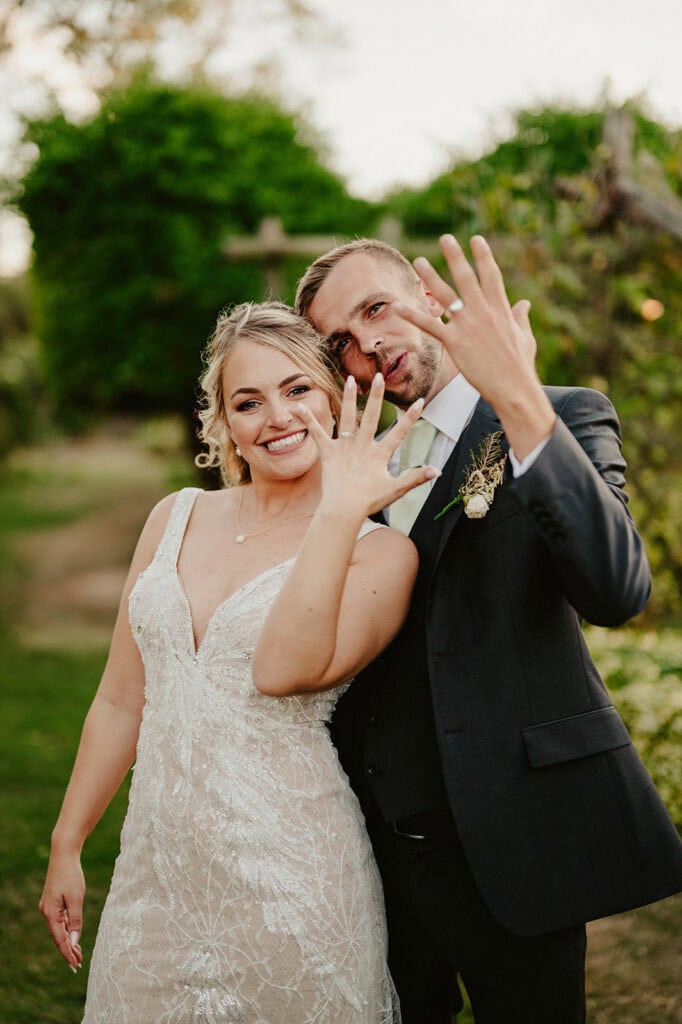 A bride and groom in wedding attire stand outdoors, smiling and showing their wedding rings to the camera.