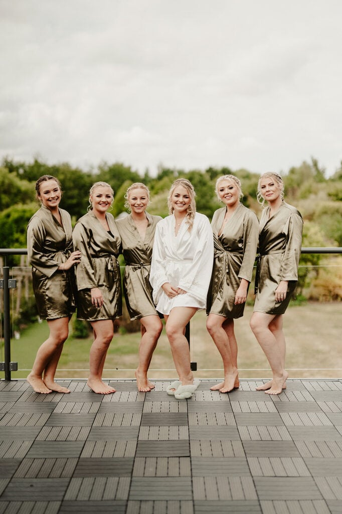 A group of six women, one in a white robe and five in matching green robes, stand on a balcony with a natural landscape in the background.
