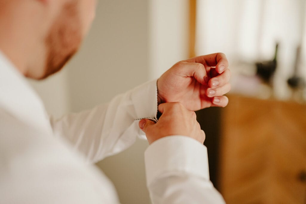 A man wearing a white dress shirt adjusts his cufflink amid the gentle glow, focusing on his hands and cuff. The background is softly blurred, reminiscent of the timeless charm found in the vines of Rochester, where elegance intertwines with mystery. Image by Pearce Wedding Photography.