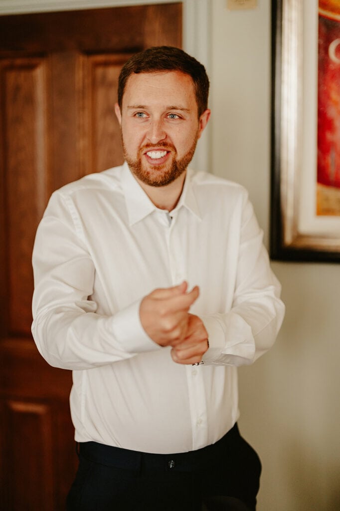 A man with a beard, wearing a white shirt, adjusts his cuffs in a well-lit room. He stands before a wooden door, where the ambiance echoes the artistry of "The Vines of Rochester," with framed artwork partially visible on the wall behind him. Image by Pearce Wedding Photography.