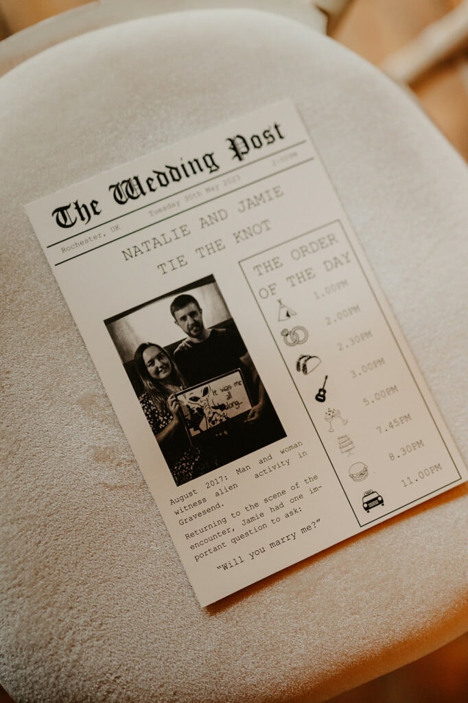 A wedding program titled "The Wedding Post" on a cushioned seat. It features a photo of a couple, order of the day schedule, and a proposal story. The event is set in Rochester, UK in August 2021.