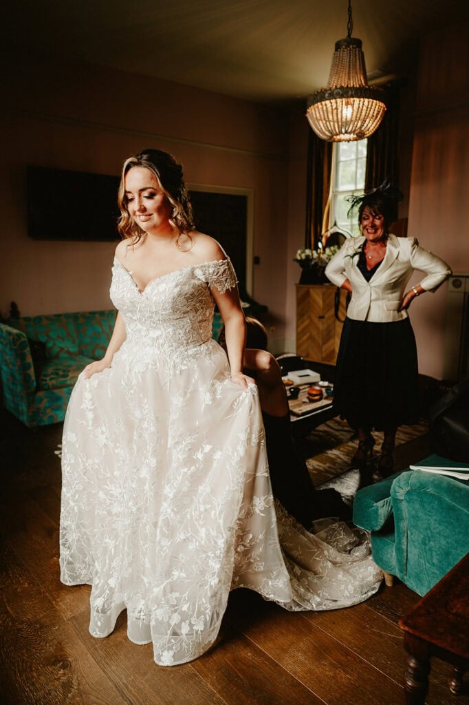 A bride in a white lace wedding dress stands in a room, lifting her gown slightly. Another woman, wearing a black and white outfit, stands nearby smiling.