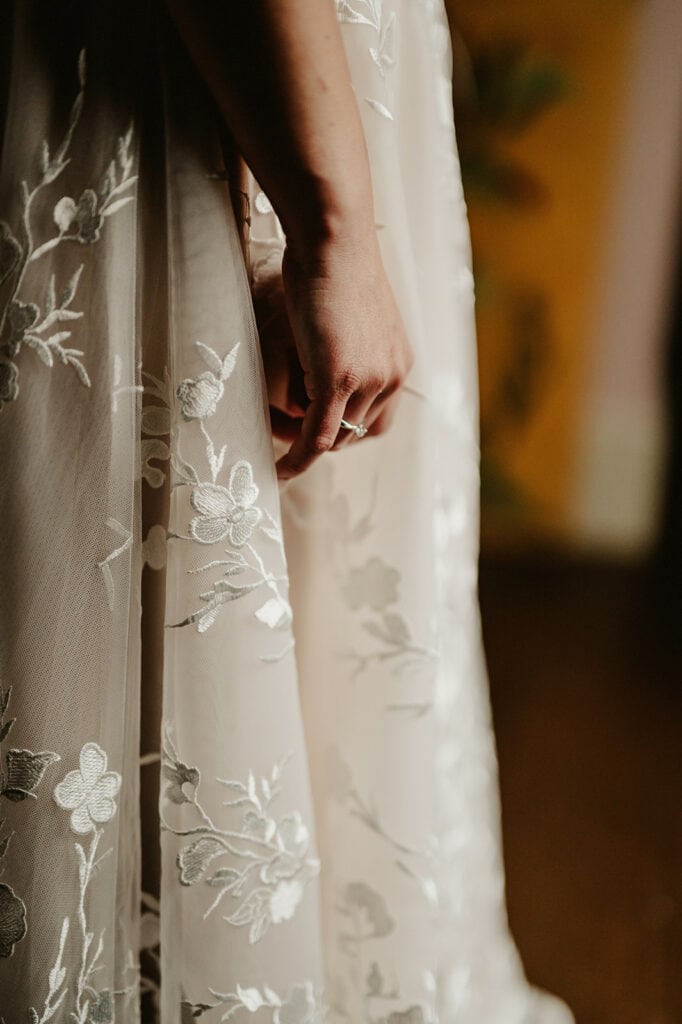 Close-up of a person wearing a delicate, floral-embroidered gown, reminiscent of the vines of Rochester. Their hand rests gently against the fabric, adorned with intricate white flowers on sheer material, capturing a soft, elegant moment. Image by Pearce Wedding Photography.