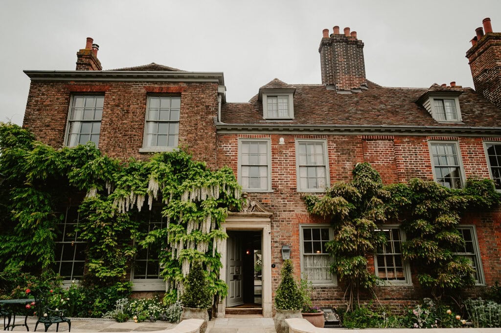 A brick house with ivy-covered walls, multiple chimneys, and tall windows. There is a front door in the middle, surrounded by greenery, giving the house a quaint and traditional appearance.