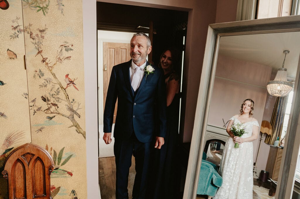 A man in a suit stands smiling in a room beside a woman behind him, while a bride in a white gown with a bouquet stands by a mirror in the background.
