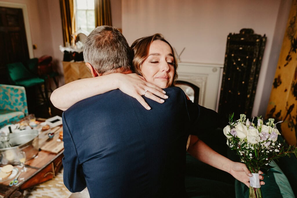 A woman holding a bouquet of flowers hugs a man in a warmly decorated room.