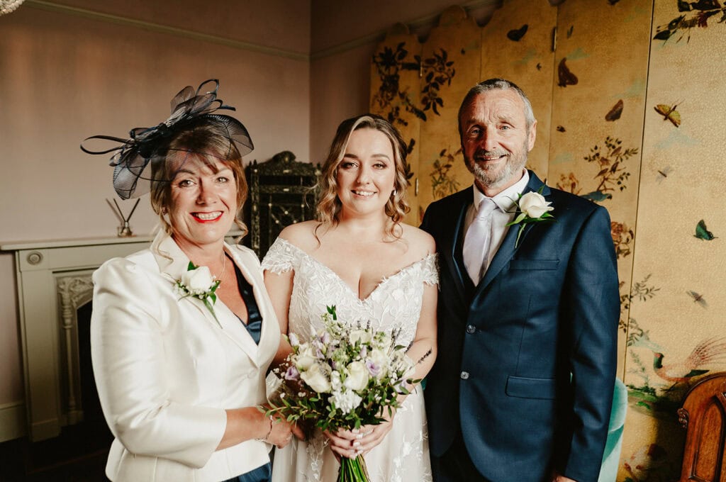 A bride in a lace gown stands between two adults, all smiling warmly in a room reminiscent of The Vines of Rochester. The woman on the left wears a white outfit with a black hat, while the man on the right sports a dark suit adorned with a white rose boutonniere. Image by Pearce Wedding Photography.