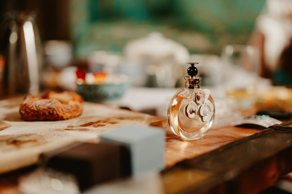 A clear, round perfume bottle with a black cap sits on a wooden table. Nearby, a pastry rests on a plate amid the warm and inviting setting of The Vines of Rochester, where blurred background elements like a black box and a cup complete the cozy scene. Image by Pearce Wedding Photography.