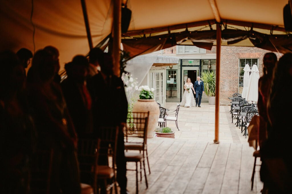 A bride, accompanied by an older man, walks down an aisle under a tent towards a group of seated guests, with a brick building visible in the background.