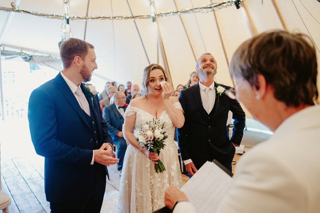 Bride and groom stand at the altar during their wedding ceremony. The bride wipes a tear, holding her bouquet, while an officiant reads from a document. Guests are seated in the background.