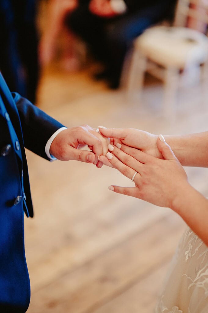 Close-up of two people holding hands, one wearing a wedding ring and the other in formal attire, on a wooden floor background.
