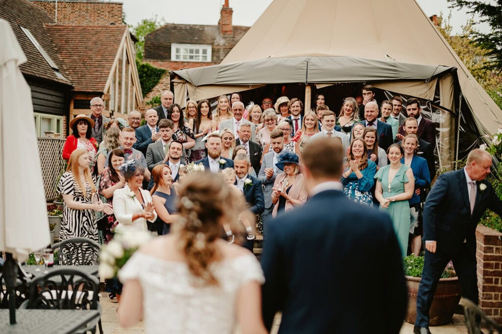 A bride and groom face a large group of seated and standing wedding guests outside a large tent at a reception. The guests are smiling and appear to be clapping and taking photos.