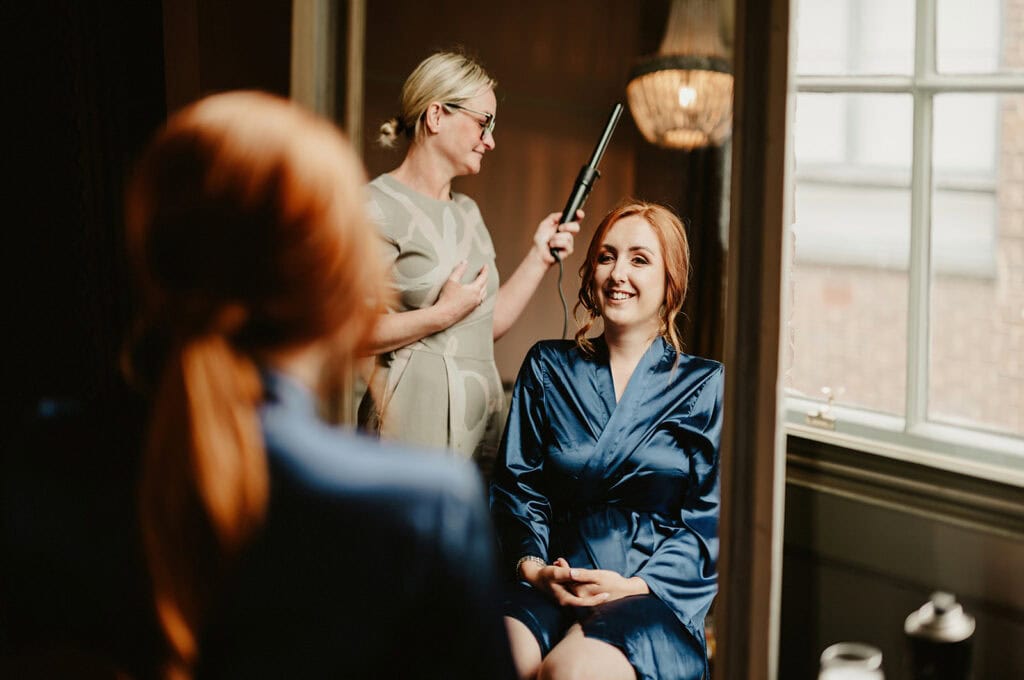 A woman in a blue robe smiles while another woman styles her hair with a curling iron. They are in a room with a large window.