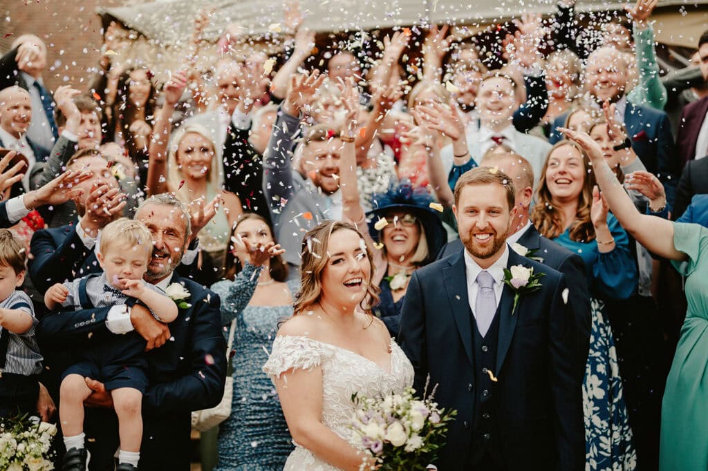 A joyful wedding scene unfolds beneath the charming arches at The Vines of Rochester, with a bride in a white dress and groom in a dark suit surrounded by well-dressed guests showering them with confetti. Smiling faces and a celebratory atmosphere highlight the happy occasion. Image by Pearce Wedding Photography.