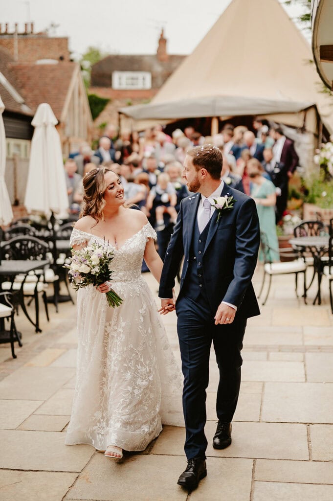 A bride and groom walk hand in hand outdoors while smiling at each other. The bride holds a bouquet, and guests are gathered in the background. Both are wearing formal wedding attire.