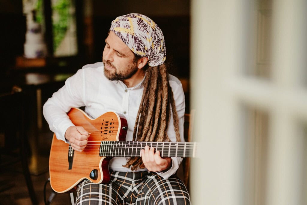 A person with long dreadlocks, wearing a patterned headscarf and plaid pants, sits on a chair playing an acoustic guitar. They are wearing a white shirt and are focused on their music, reminiscent of the soulful vibes found in The Vines of Rochester. The setting appears to be indoors. Image by Pearce Wedding Photography.
