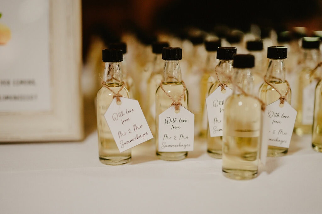 Small bottles with handwritten tags, "With love from Mr. & Mrs. Sommaburangas," are elegantly arranged on a white table at The Vines of Rochester. Each bottle, topped with a cork stopper and filled with light-colored liquid, hints at being charming party favors or thoughtful gifts. Image by Pearce Wedding Photography.