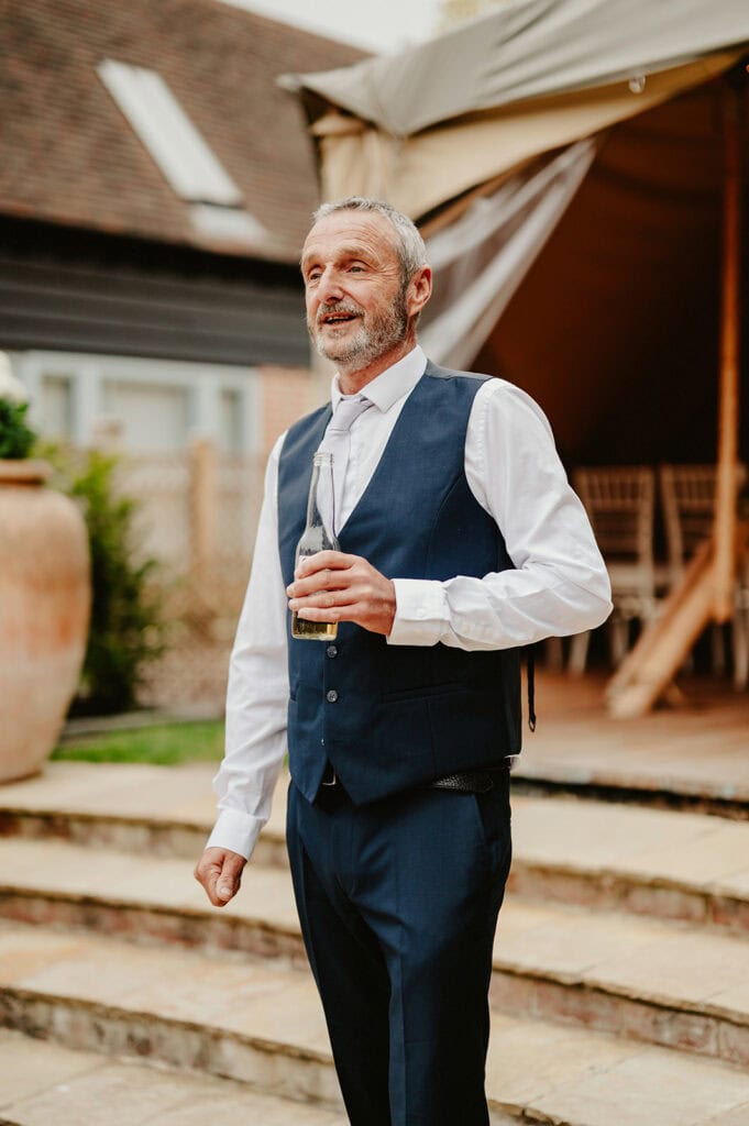 A man in a navy suit vest and white shirt holds a drink while standing outside near a tent at a social event.