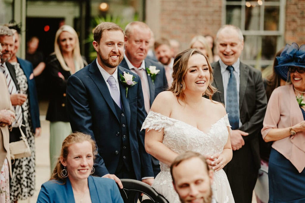 A bride and groom, dressed formally, stand and smile among a crowd of people at a wedding reception. The bride is in a white off-shoulder dress, and the groom is in a navy suit with a lavender tie.