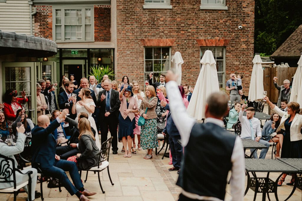 A group of people dressed in formal attire gather outdoors in a courtyard, raising glasses in a toast, with a brick building in the background.