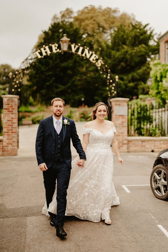 A bride and groom hold hands while walking in front of a brick gate with "The Vines" sign overhead. Both are smiling. The bride wears a white gown; the groom wears a dark suit and tie.