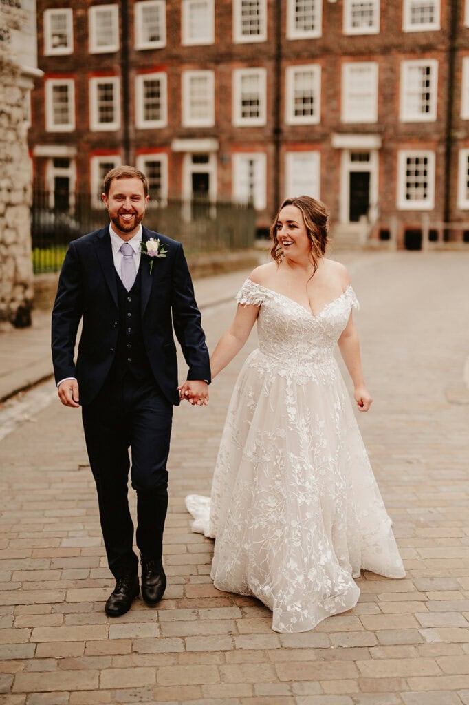 A bride in a lace wedding dress and a groom in a navy suit walk hand-in-hand along the cobblestone street. They are smiling joyfully against the backdrop of the historic building, where the vines of Rochester gently cascade down its aged facade. Image by Pearce Wedding Photography.
