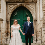A couple in wedding attire stands holding hands before the large, ornate green wooden door of The Vines of Rochester, set in a stone wall. The bride glows in her white lace dress, while the groom looks dapper in his navy blue suit with a boutonniere. Both are smiling warmly at the camera. Image by Pearce Wedding Photography.