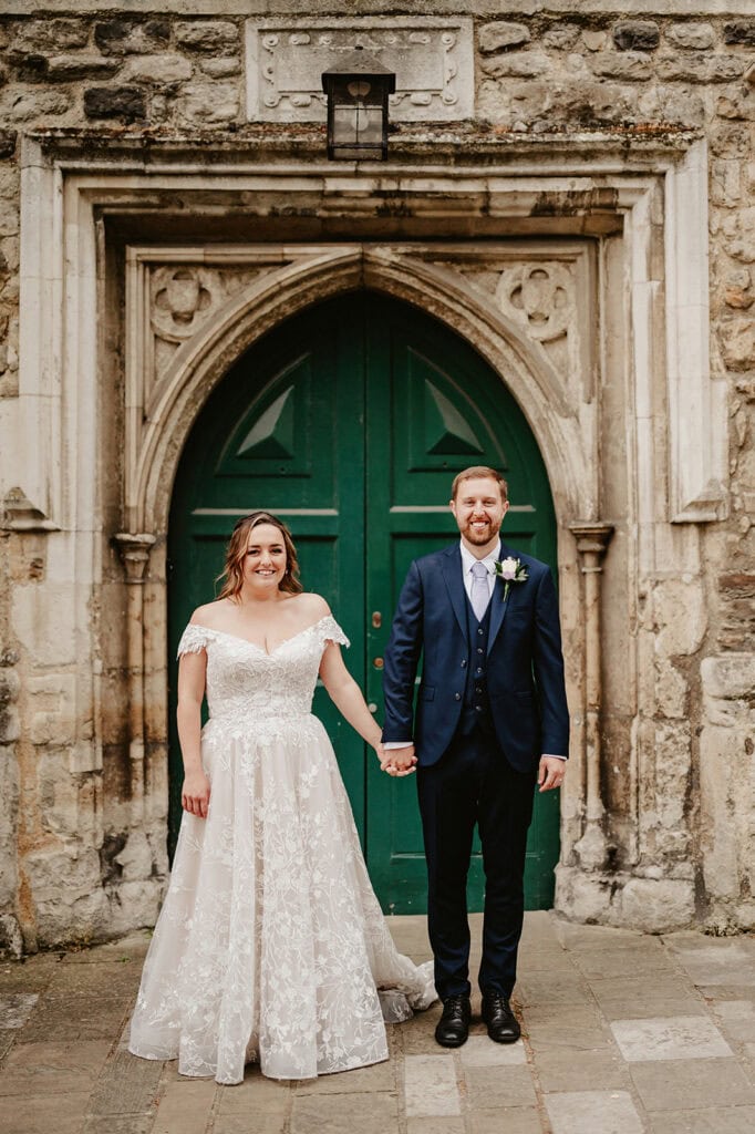 A couple in wedding attire stands holding hands before the large, ornate green wooden door of The Vines of Rochester, set in a stone wall. The bride glows in her white lace dress, while the groom looks dapper in his navy blue suit with a boutonniere. Both are smiling warmly at the camera. Image by Pearce Wedding Photography.