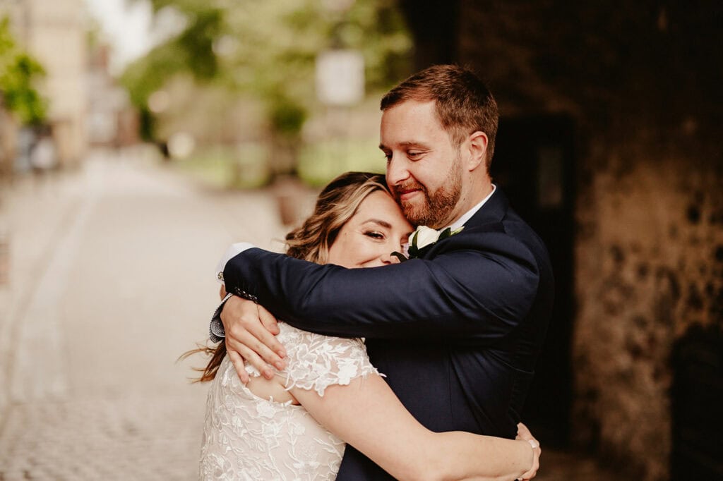 A couple in formal attire embraces lovingly outdoors. The man, wearing a navy suit, holds the woman, dressed in a white lace outfit, as she rests her head on his shoulder. The background is blurred.