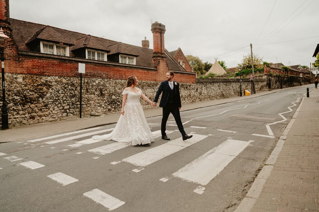 A bride and groom walk hand in hand across a crosswalk on a street lined with brick and stone buildings. Under the cloudy Rochester sky, which enhances the moody backdrop, she wears a long white gown while he dons a dark suit, mirroring the city's charming contrast. Image by Pearce Wedding Photography.