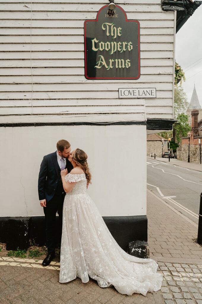 A couple in wedding attire shares a kiss on a street corner, beneath signs reading "The Coopers Arms" and "Love Lane." The bride wears a lace gown, the groom dons a suit. In the background, The Vines of Rochester and a charming church whisper timeless romance among the trees. Image by Pearce Wedding Photography.