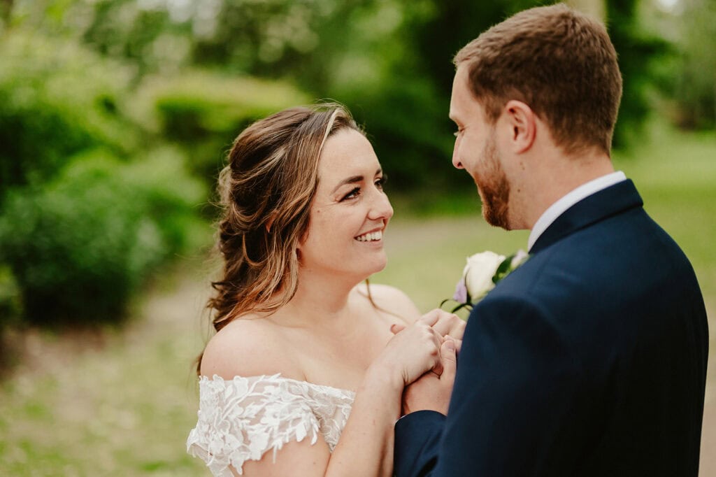 A couple stands outside, smiling at each other. The woman is wearing an off-shoulder white dress, and the man is in a navy suit. They hold hands against a backdrop of lush greenery, reminiscent of the vines of Rochester weaving through their perfect moment. Image by Pearce Wedding Photography.