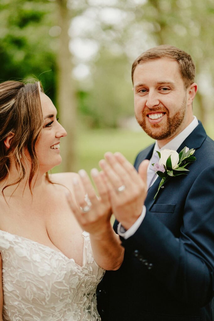 A joyful couple on their wedding day, standing outdoors at The Vines of Rochester. The bride, in a white lace dress, extends her hand with a wedding ring, sharing a smile with the groom in a dark suit and white boutonniere. Lush greenery surrounds them. Image by Pearce Wedding Photography.