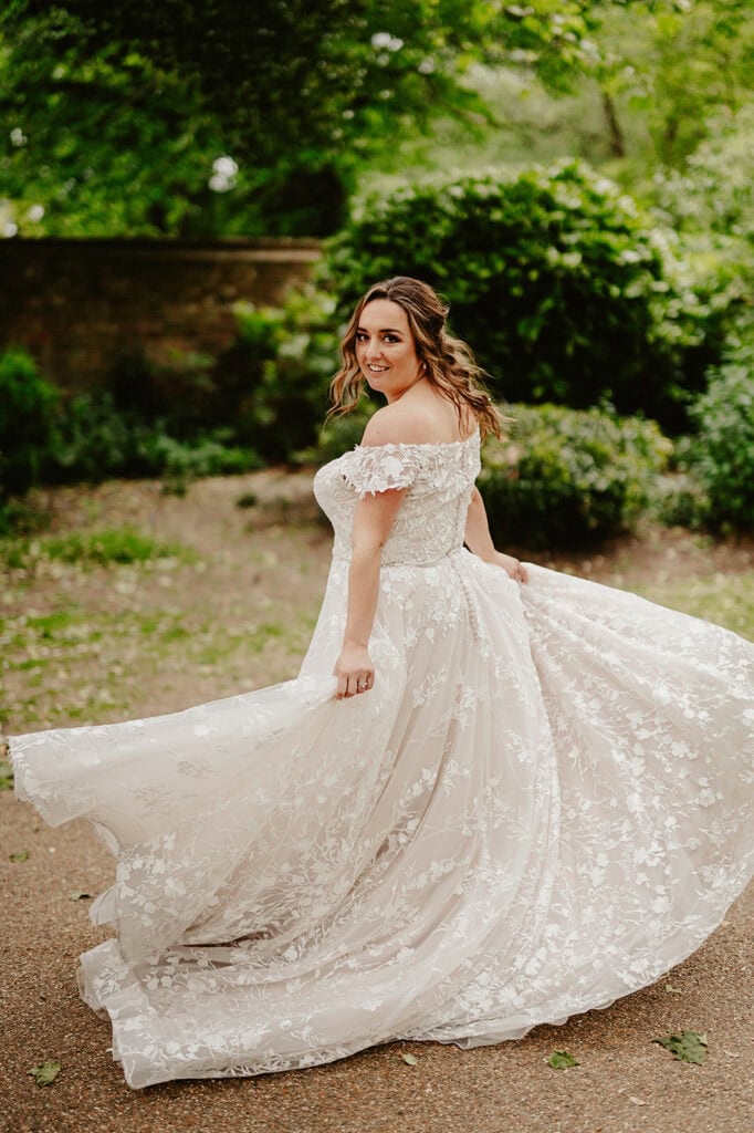 A woman in a white, off-the-shoulder wedding dress twirls outdoors, surrounded by greenery.