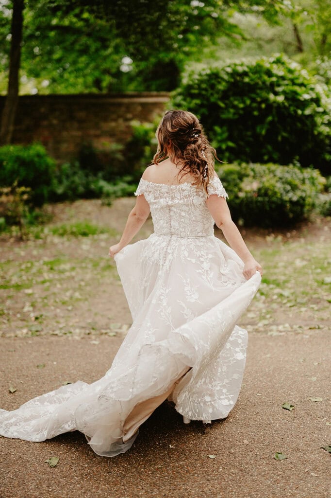 A woman in a white wedding dress walks away on a path, holding the sides of her gown. She has long wavy hair and is surrounded by trees and greenery.
