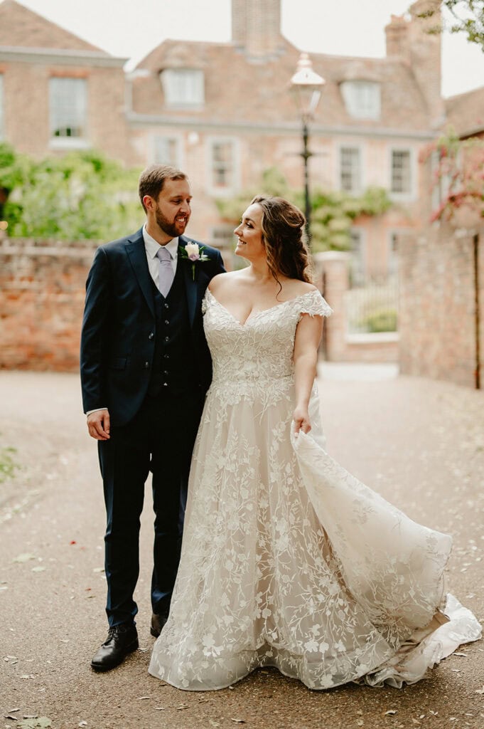 A bride in a white lace gown and a groom in a dark suit stand closely together outdoors in front of a brick building.