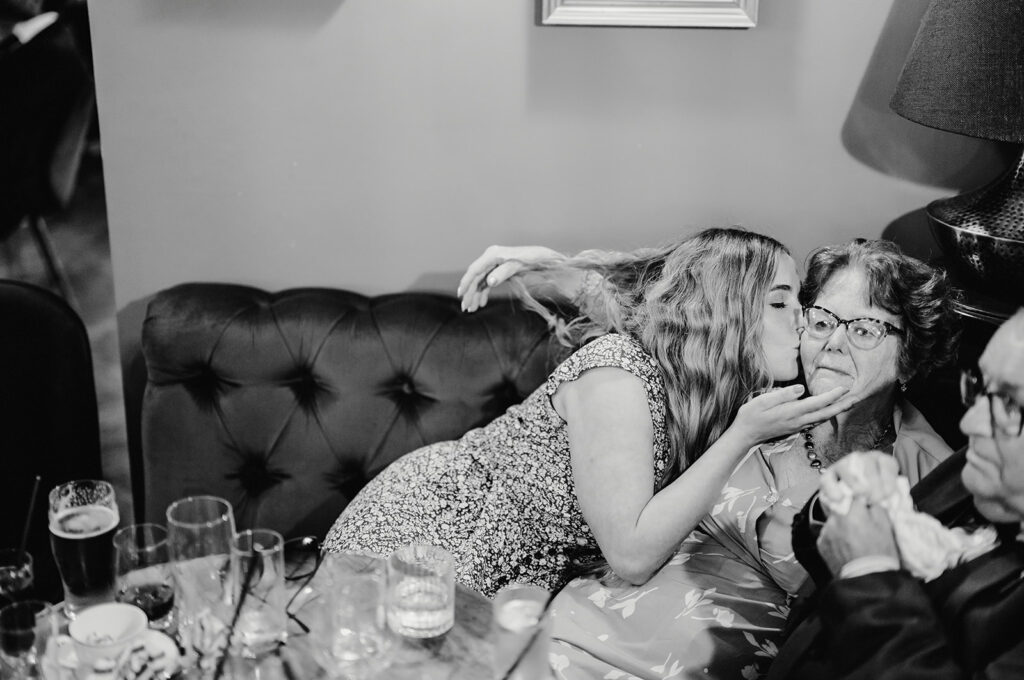 A young woman kisses an older woman on the cheek while seated together on a couch. The older woman appears surprised. Drinks and glasses are on a table in the foreground. Black and white image.