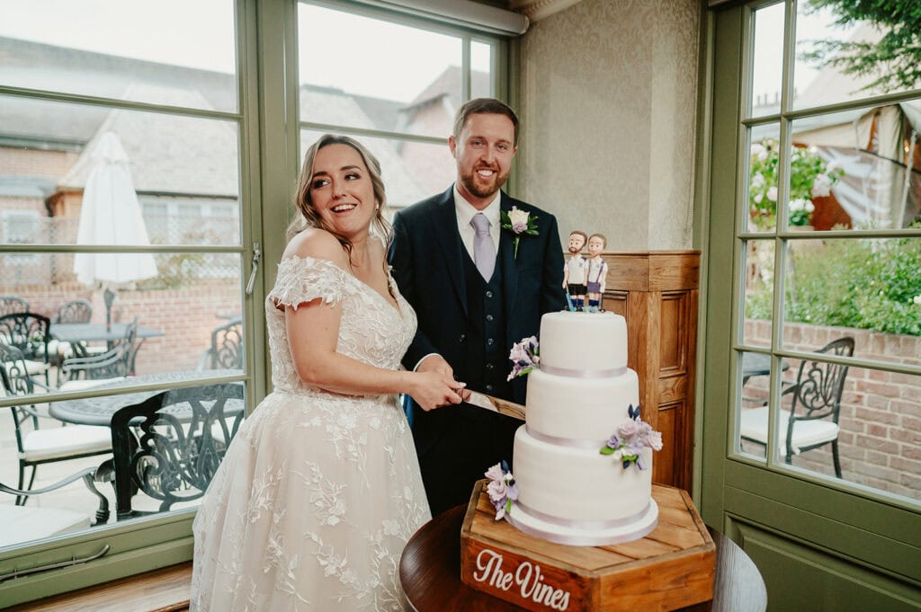 A bride and groom are cutting a three-tiered wedding cake. The cake topper features figures resembling the couple. They are smiling and standing close together. The setting appears to be an indoor venue with large windows.