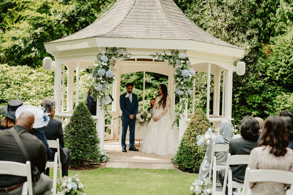 A couple stands under a flower-adorned gazebo in Maidstone, holding hands during their wedding ceremony. The groom wears a blue suit, and the bride dazzles in a white gown. Guests sit on white chairs, witnessing the event in a lush garden setting as the photographer captures every precious moment. Image by Pearce Wedding Photography.