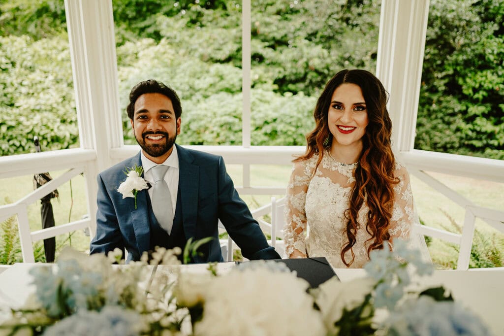 A man in a blue suit and a woman in a white lace dress sit together at a table adorned with blue and white flowers, with greenery visible in the background.