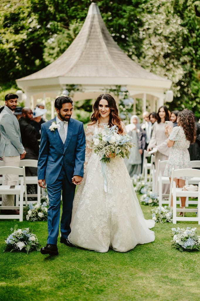 A bride and groom walk down an outdoor aisle together, smiling and holding hands. They are surrounded by seated and standing guests, with a white gazebo in the background.