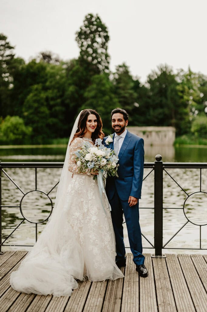 A couple in wedding attire poses together on a wooden deck by a lake, surrounded by greenery. The bride holds a bouquet of flowers.