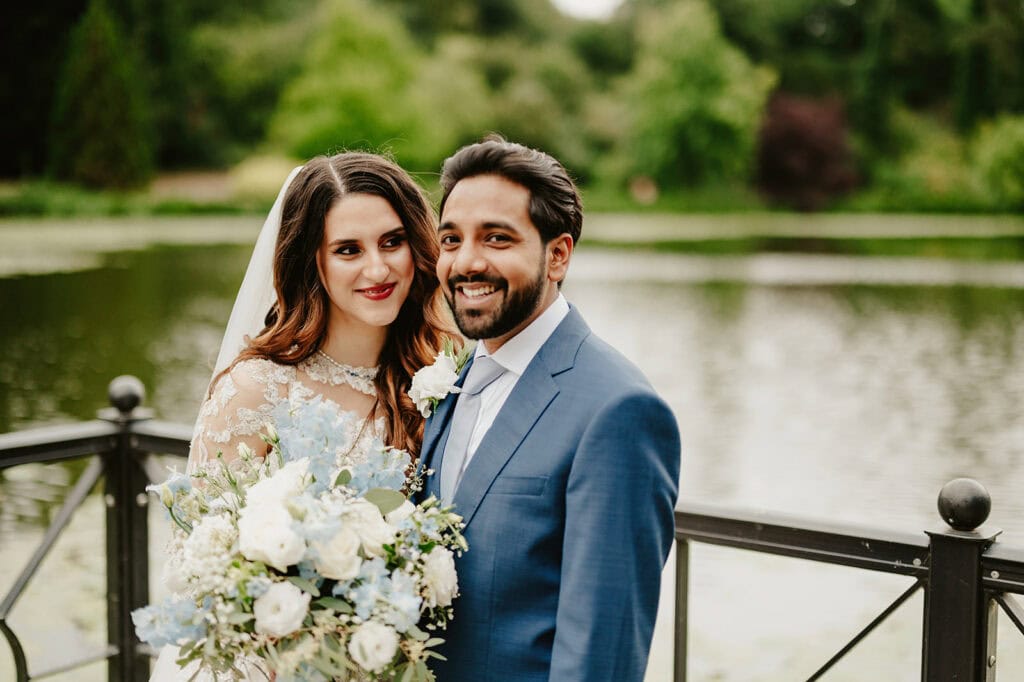 A smiling bride and groom stand together outdoors by a serene lake. The bride holds a bouquet of white and light blue flowers and wears a lace wedding dress with a veil, while the groom is dressed in a blue suit and tie. Green trees are visible in the background, captured beautifully by their Maidstone wedding photographer. Image by Pearce Wedding Photography.